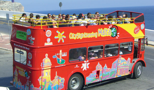 A bright red Malta Bus glides through the city, delighting its passengers as they soak in the sights and sounds of their journey.