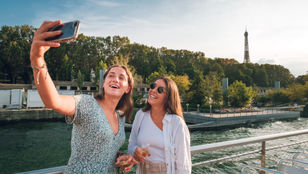 Two friends are trying to catch the perfect moment by smiling and clicking a lovely selfie against the background of the Seine River Paris.