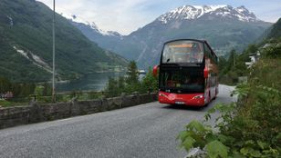 A striking red hop-on hop-off bus cruising through the streets of Geiranger, framed by majestic mountains, creating a stunning scenic experience.