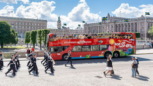 Red Sightseeing Stockholm Hop-On, Hop-Off Bus glides through Stockholm's streets, accompanied by a vibrant parade on the side.