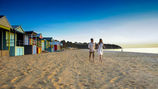 A couple relishing their time at a Melbourne beach, with colorful hut-shaped houses dotting the shoreline in the background.