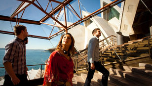 A group of excited tourists climb the stairs of Sydney's famous Opera House, while admiring its beauty and architecture.