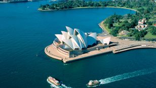 From above, a cruise ship glides on the sea, framed by Sydney's iconic Opera House, a picturesque blend of nature and culture.